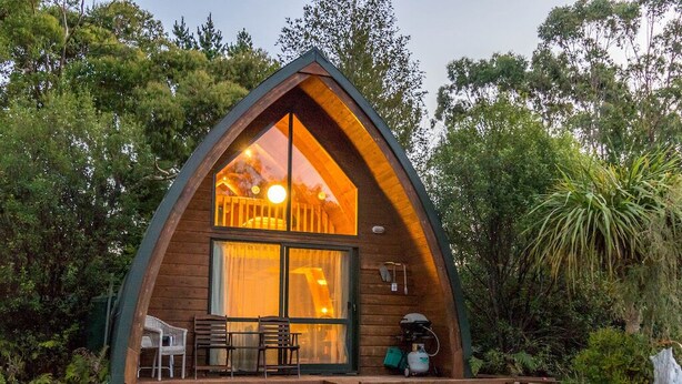 Un chalet confortable en forme de « A », avec une structure en bois, une grande porte et de grandes fenêtres en verre, le tout entouré d’une végétation luxuriante.