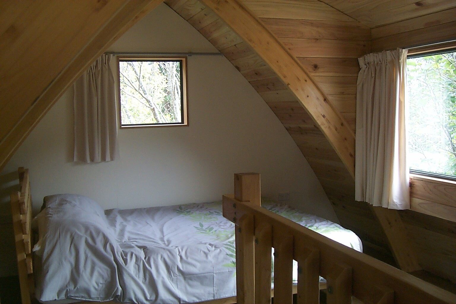 Une petite chambre mansardée, chaleureuse et accueillante, avec un lit simple, des poutres en bois et deux fenêtres dotées de rideaux.