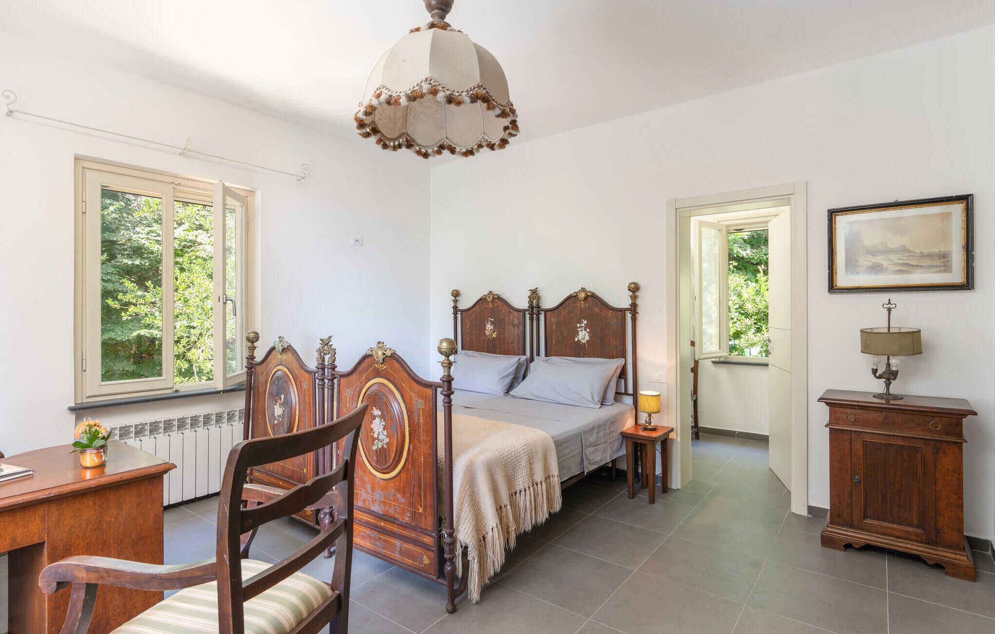 Ein Schlafzimmer mit einem großen Bett, einem Schreibtisch, einem Stuhl, einem Nachttisch mit Lampe und einem Schrank. Zwei Fenster und eine Tür zu einem anderen Raum sind vorhanden.