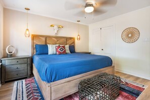 Quarto com cabeceira de madeira, cama com roupa de cama azul, duas luminárias de apoio, espelho redondo, cesta de fibra natural e tapete estampado.