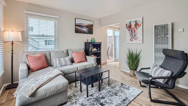 A modern living room with a grey sectional sofa, a black coffee table, and a grey armchair. There is a bookshelf with books and decorative items, a floor lamp, and a large window with blinds. - OB Cottage w outdoor patio / 3 min to the beach   (San Diego)