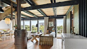 Ein rustikales Interieur mit Holzbalkendecke, Kamin aus Stein, einem Essbereich mit Holztisch und -stühlen sowie Blick ins Freie durch große Fenster.