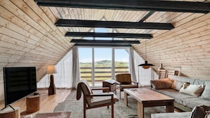 Ein gemütliches Dachzimmer mit Holzbalkendecke, einem großen Fenster, einem Fernseher, einer Couch, einem Sessel, einem Tisch und einer Lampe.