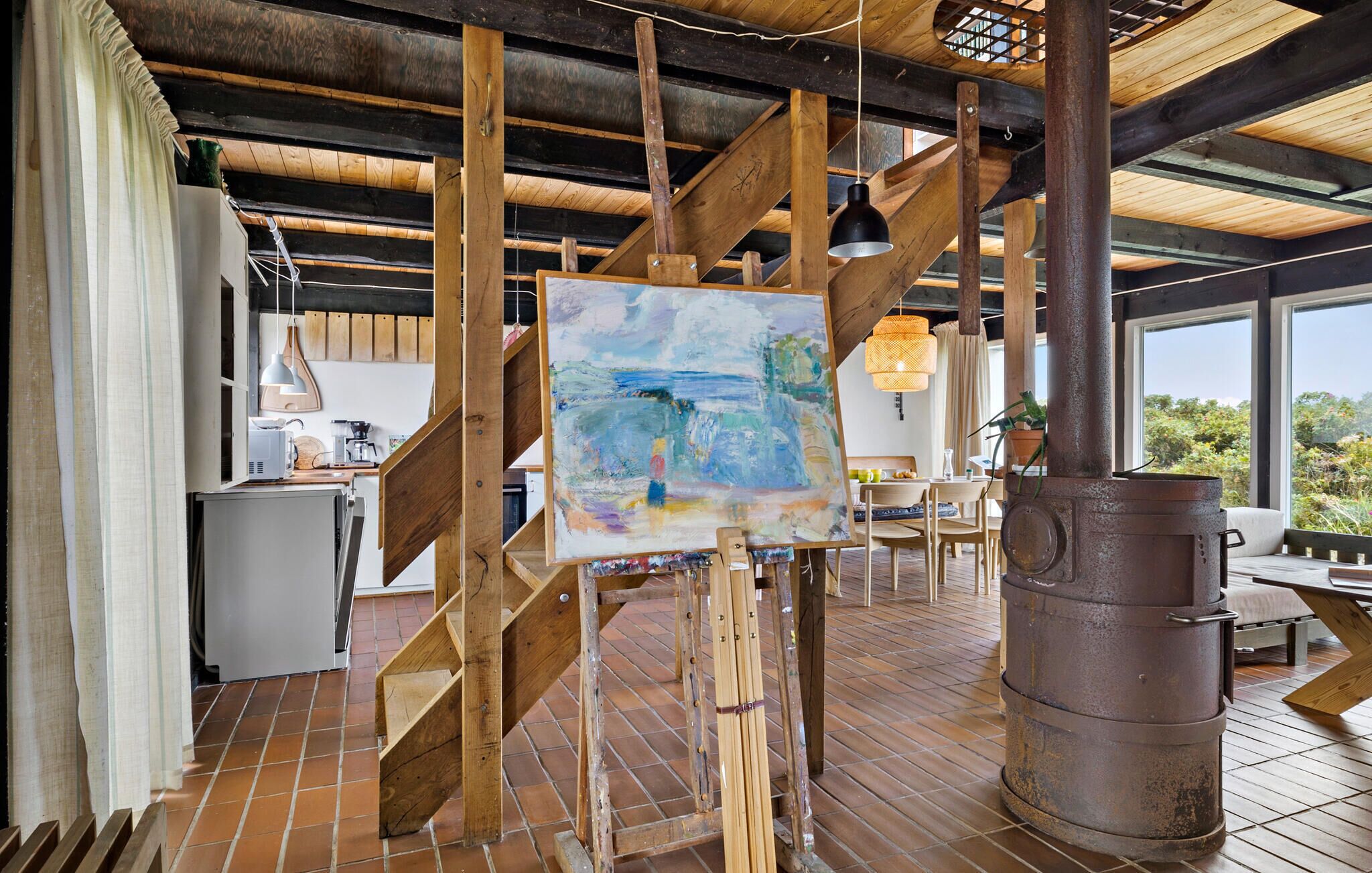 Un intérieur rustique avec des poutres apparentes, un grand tableau posé sur un chevalet en bois et un poêle rustique.