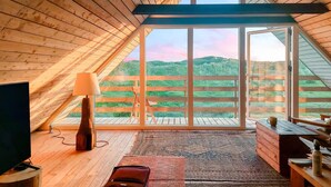 Ein gemütliches Zimmer mit großem Fenster, Holzboden, Fernseher, Lampe und kleinem Tisch.