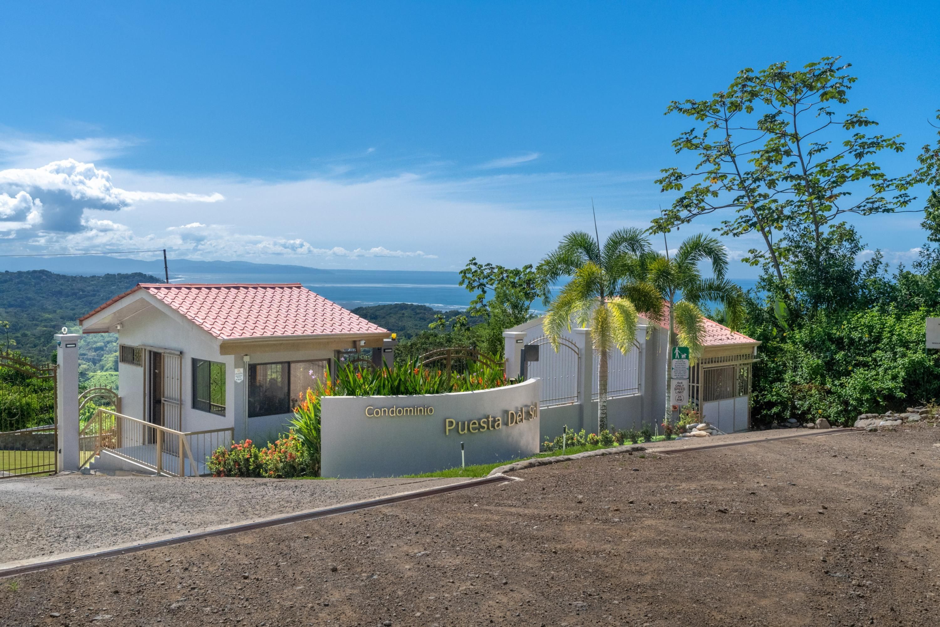 A gated entrance with a sign reading 'Condominio Puesta Del Sol,' a small building with a red-tiled roof, and a view of the ocean and hills in the background.