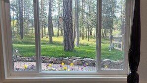 A room with a large window overlooking a grassy area with trees and a stone wall. - Montana Forest Retreat, Easy Walk to Flathead Lake, 1.5 Acres Fenced for Dogs! (Polson)