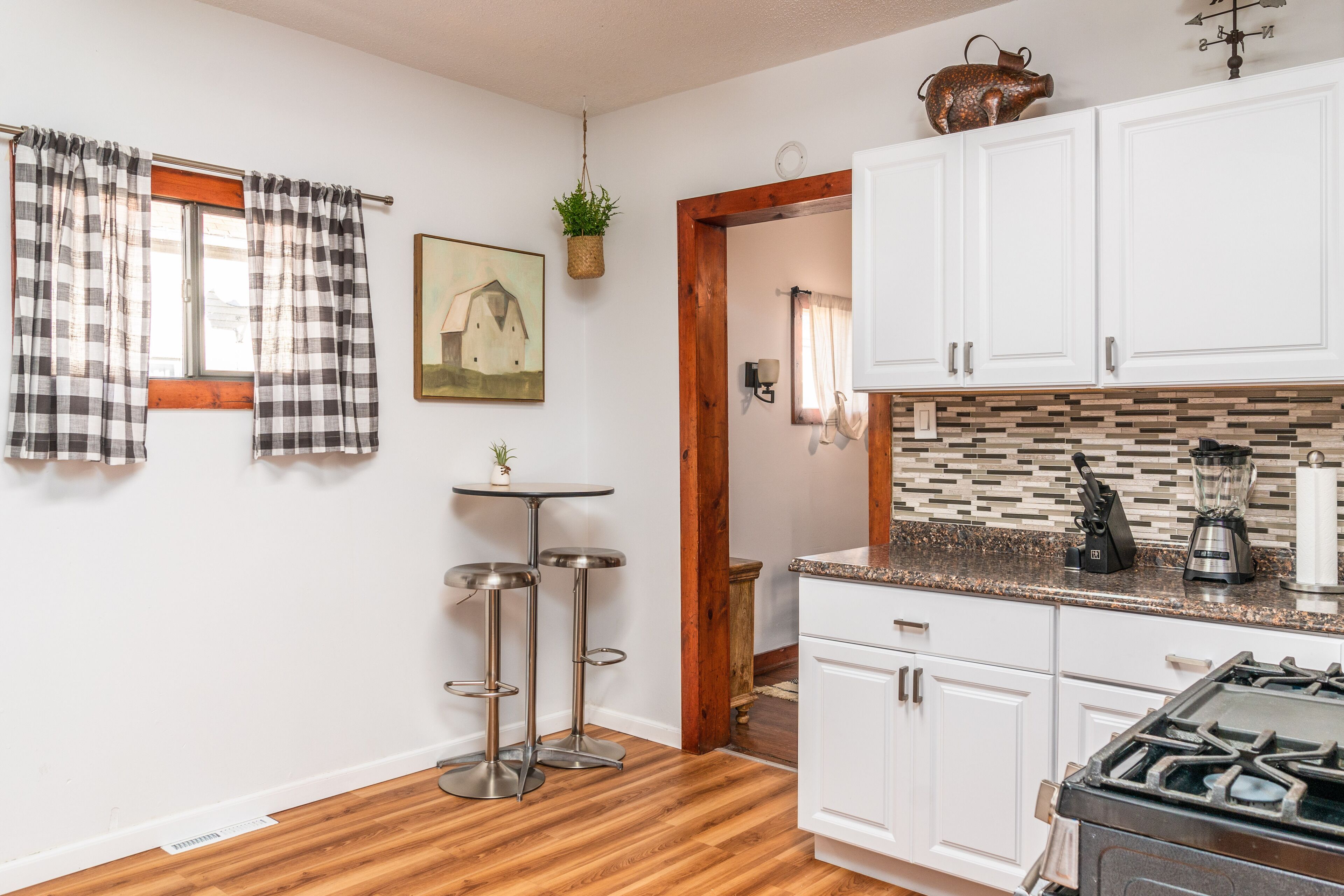 A kitchen with white cabinets, a stove, a countertop with a blender, and a window with plaid curtains.