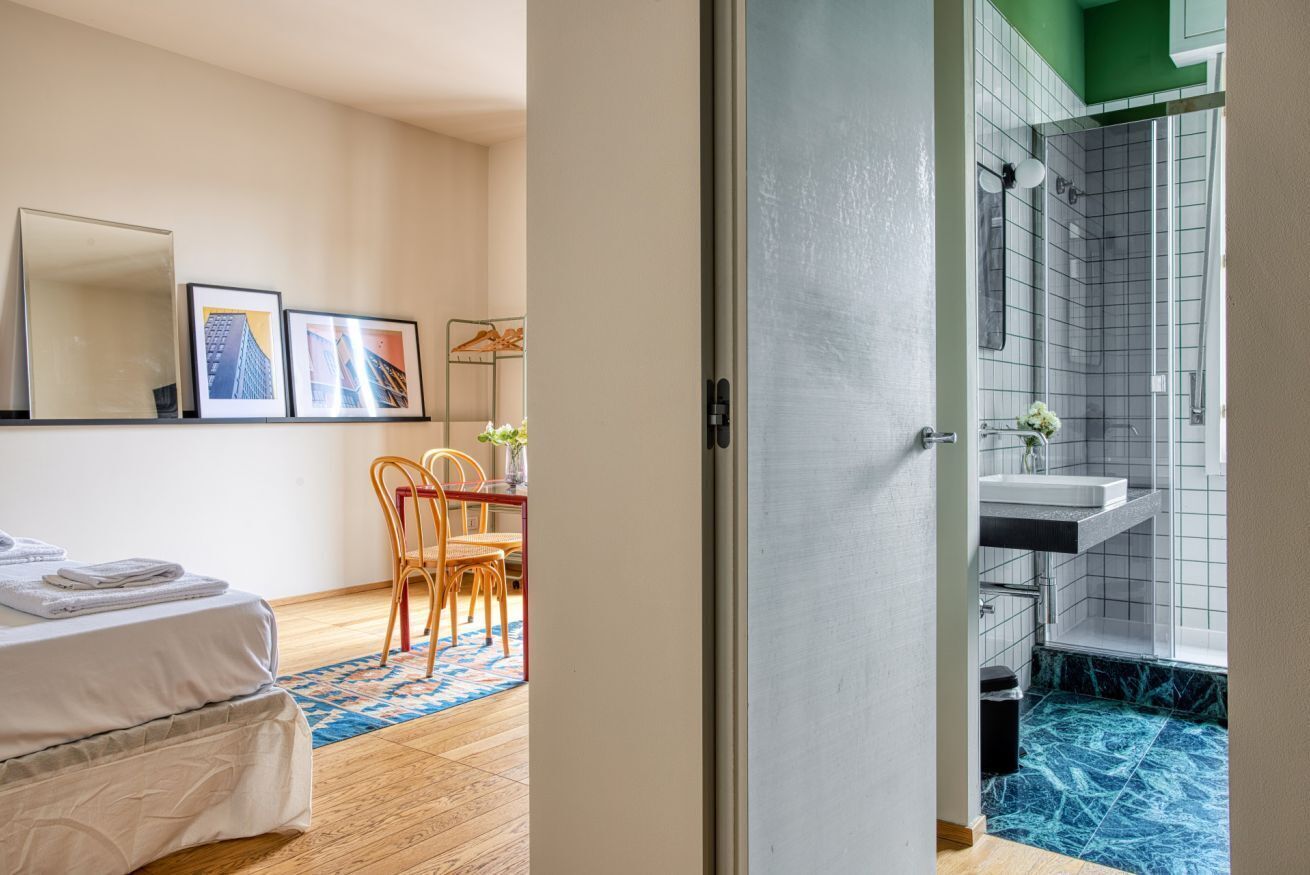 A hotel room with a bed, a dining area with chairs, a table, and a view of the bathroom with a shower and sink.