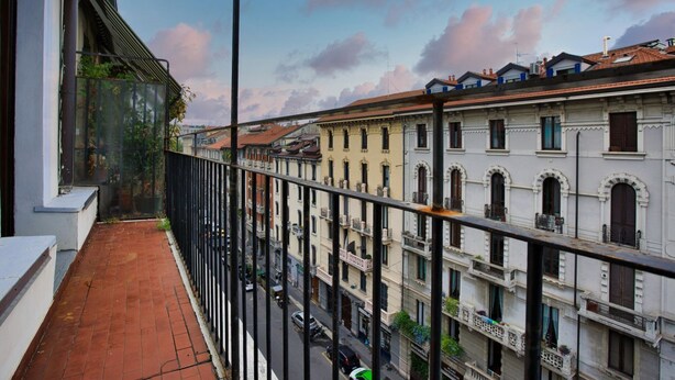 Ein Balkon mit rotem Backsteinboden, der auf eine Straße mit historischen Gebäuden und parkenden Autos blickt.