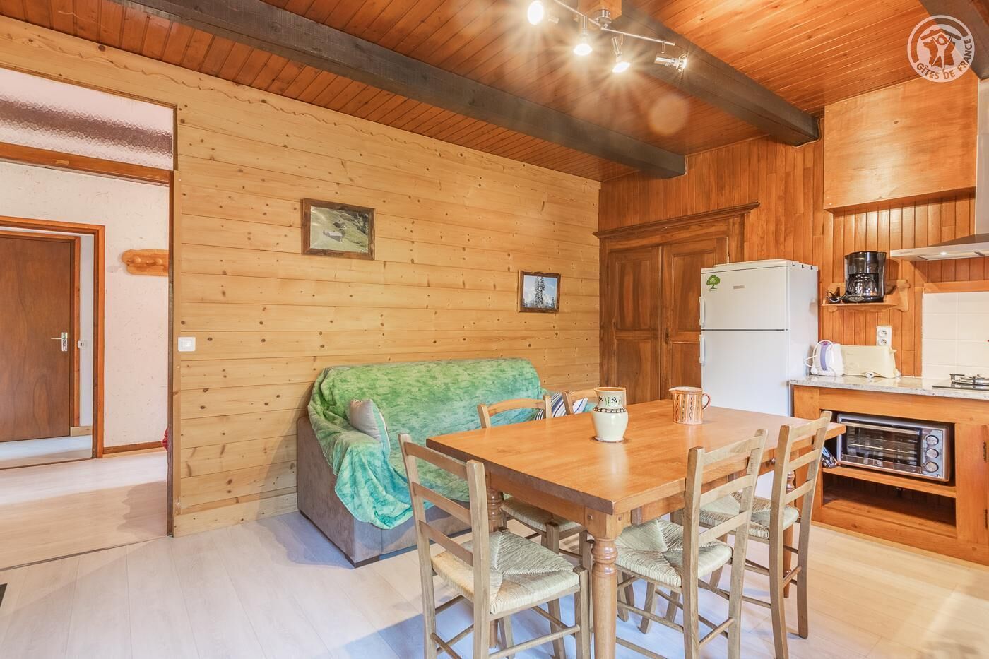 L’intérieur chaleureux d’un chalet avec une table à manger en bois, des chaises, un canapé recouvert d’une couverture verte, un coin cuisine équipé d’un réfrigérateur, d’une machine à café et d’un grille-pain.