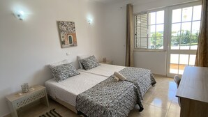 A bedroom with a bed, a bedside table, a rug, a window with curtains, and a wall-mounted picture. - Vila Olival, Bbq, 2 Quartos, Espaço Exterior (Albufeira)