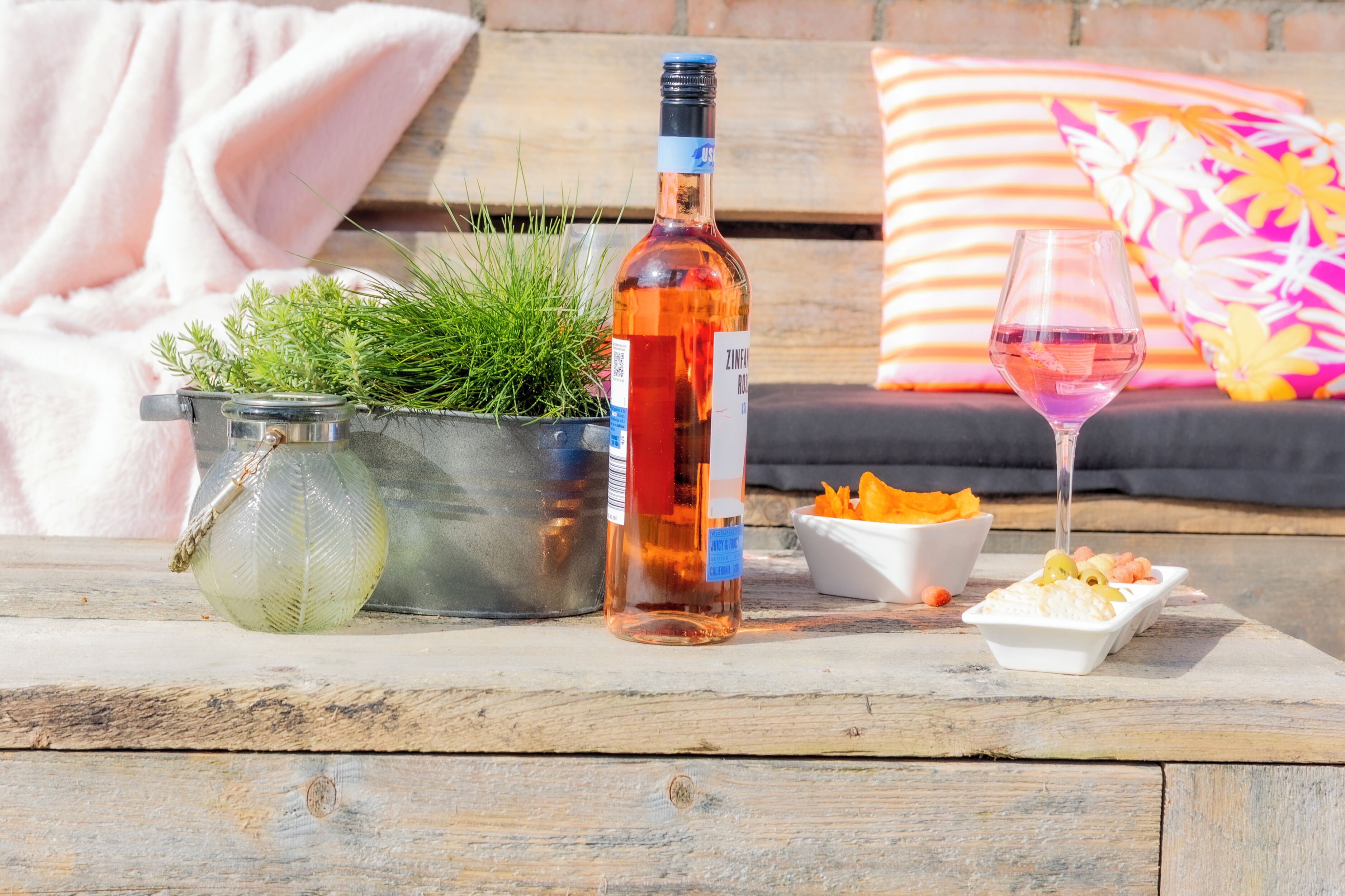 Una botella de vino Zinfandel, una copa de vino rosado, un colador metálico con plantas verdes y un cuenco blanco con comida sobre una mesa de madera.