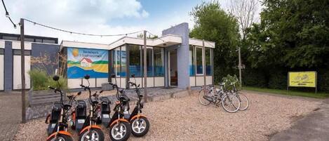 Een gebouw met een modern ontwerp, met een glazen gevel en een muurschildering. Drie elektrische scooters en twee fietsen staan er buiten geparkeerd.