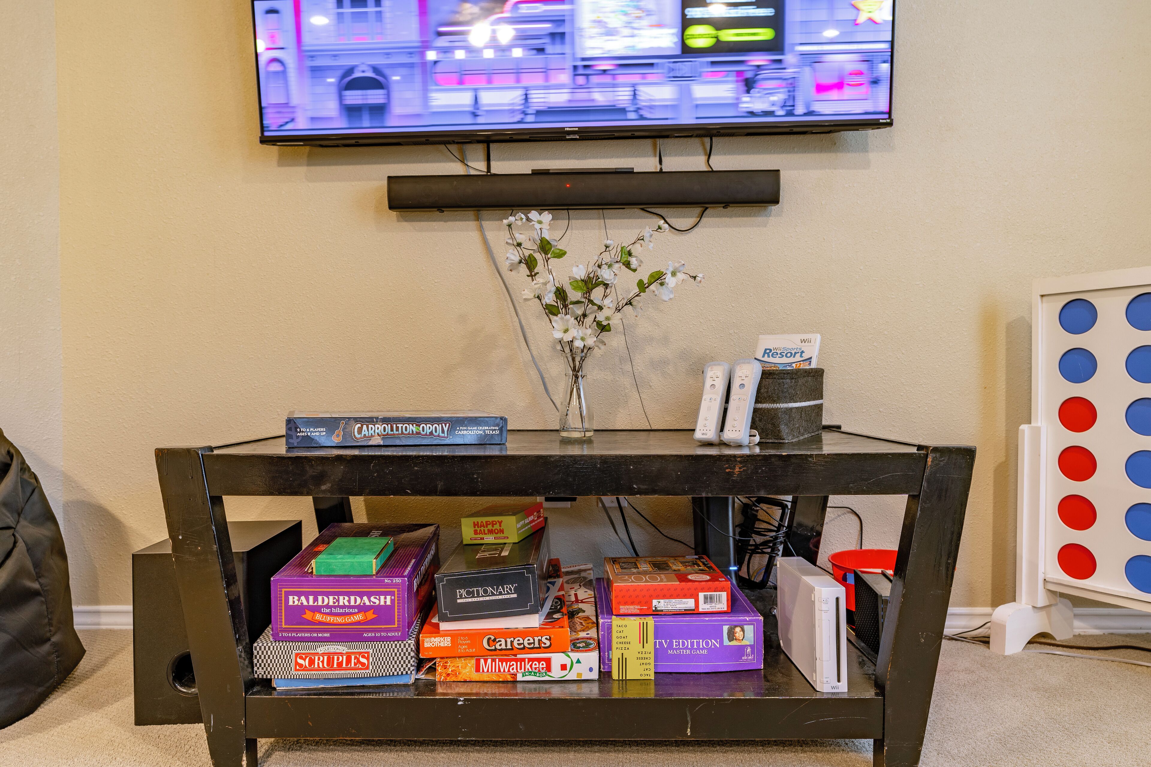 Una consola de videojuegos sobre una estantería, un televisor montado en la pared y varios juegos de mesa.