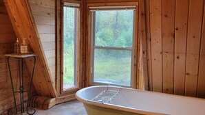 Interior d'una cabana de fusta amb una banyera, dues finestres i una taula petita amb productes de tocador.