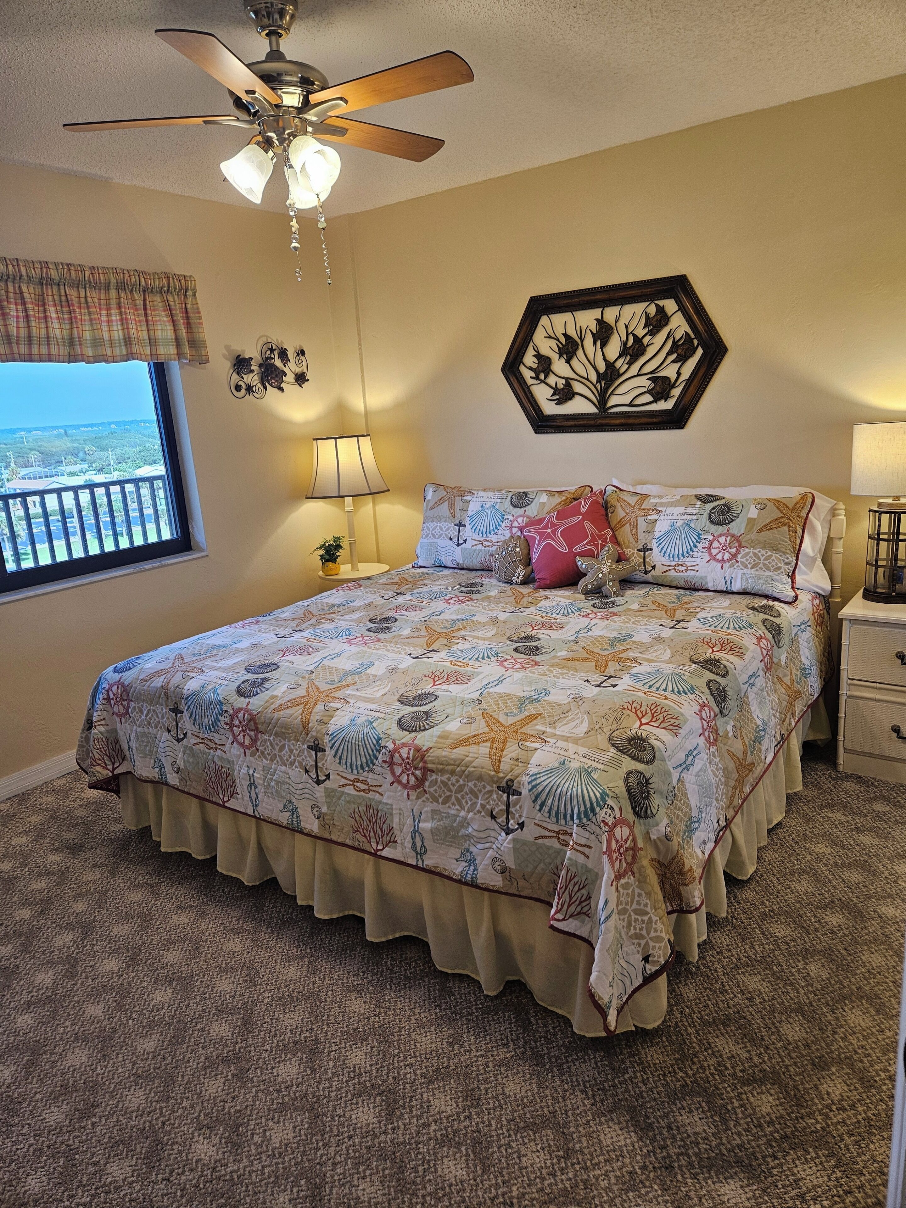 A bedroom with a bed, a ceiling fan, a window with a view, a nightstand, and a wall decoration.