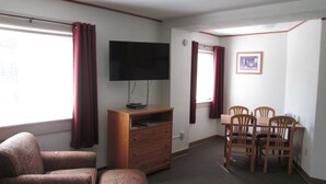 Quarto com televisão de tela plana em suporte de madeira, mesa de jantar com quatro cadeiras, sofá estampado e janela com cortinas.