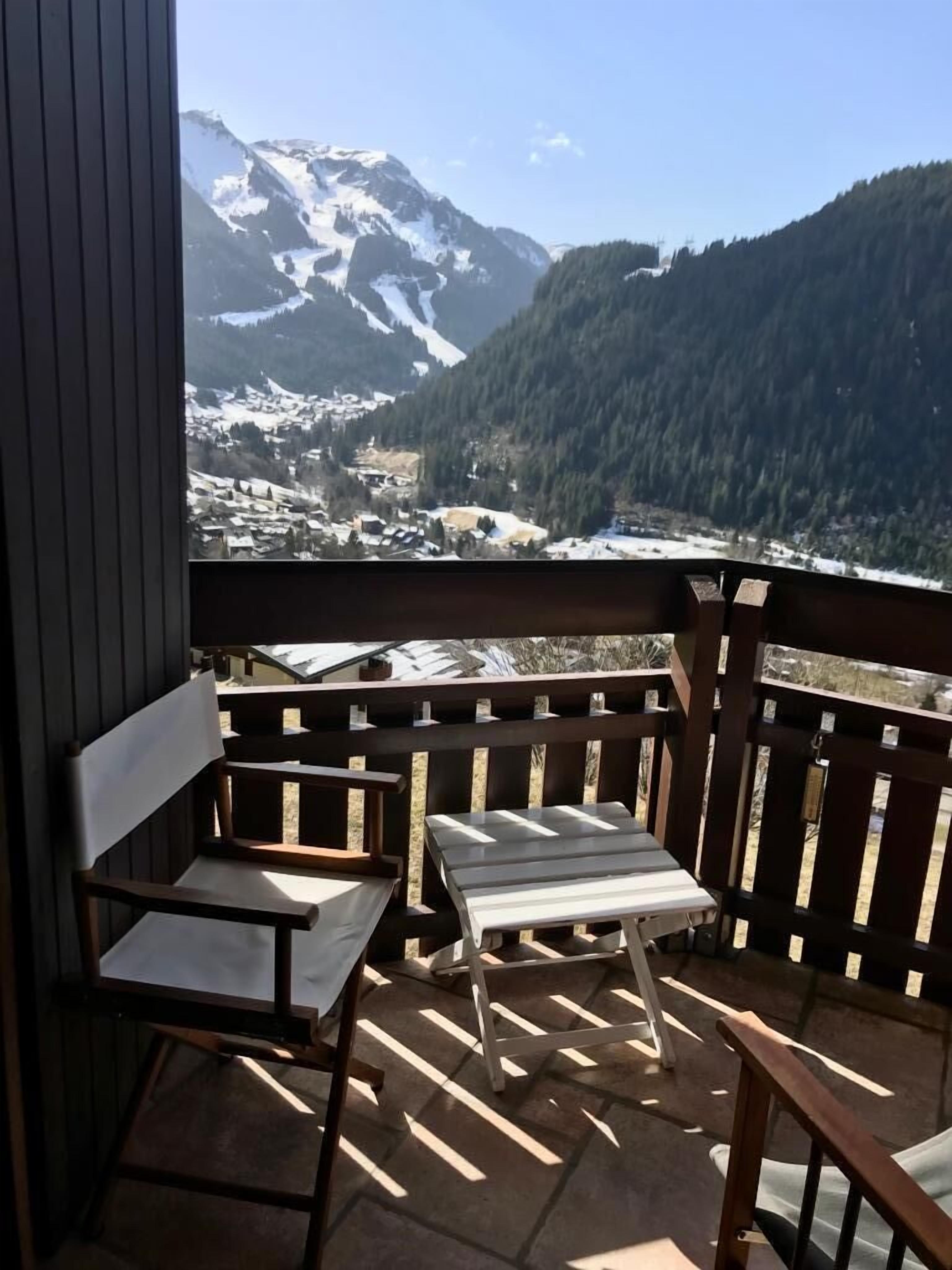 A balcony with a wooden bench and a table, overlooking a mountainous landscape with snow-covered peaks and a valley below.
