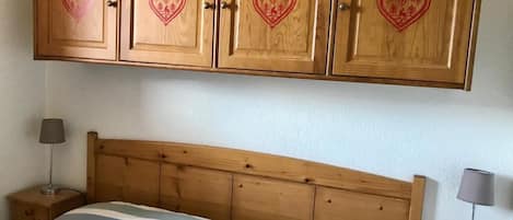 A bedroom with a wooden headboard, a bed with striped bedding, and a nightstand with a lamp. Above the bed, there is a wooden cabinet with heart designs.
