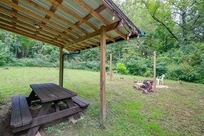 Ein Holztisch mit Picknickplatz unter einem Metalldach, umgeben von einer Rasenfläche mit Bäumen und einem Holzstapel.
