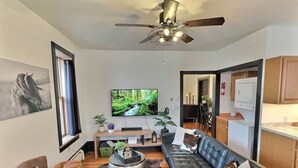 A modern living room with a leather sofa, a television, a ceiling fan, and a kitchen area visible through an open doorway. - Fully-furnished Modern Apartment / Executive Suite in Heart of Town
 (Big Timber)