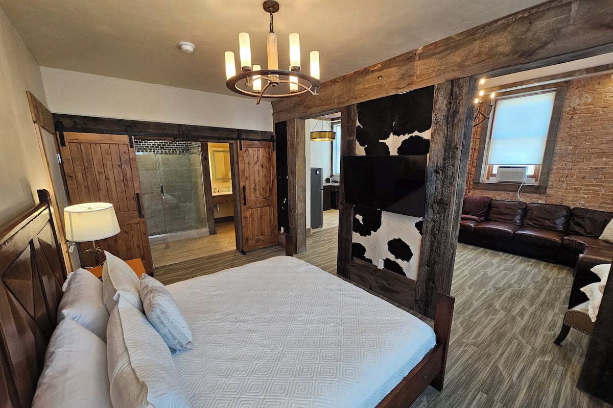 A bedroom with a large bed, a wooden headboard, a chandelier, a cowhide wall panel, a leather sofa, and a bathroom with a glass shower enclosure.