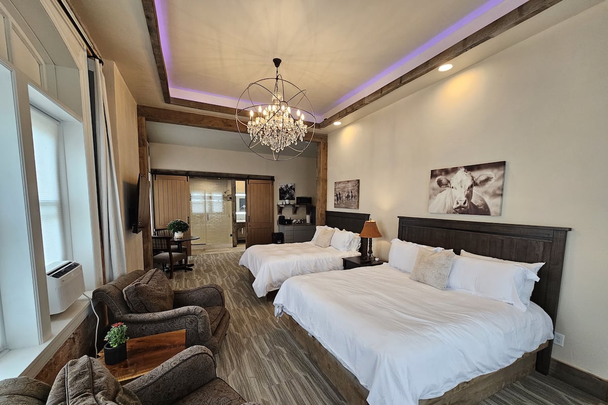 A hotel room with two beds, a chandelier, a desk with a lamp, a sofa, and a large window.