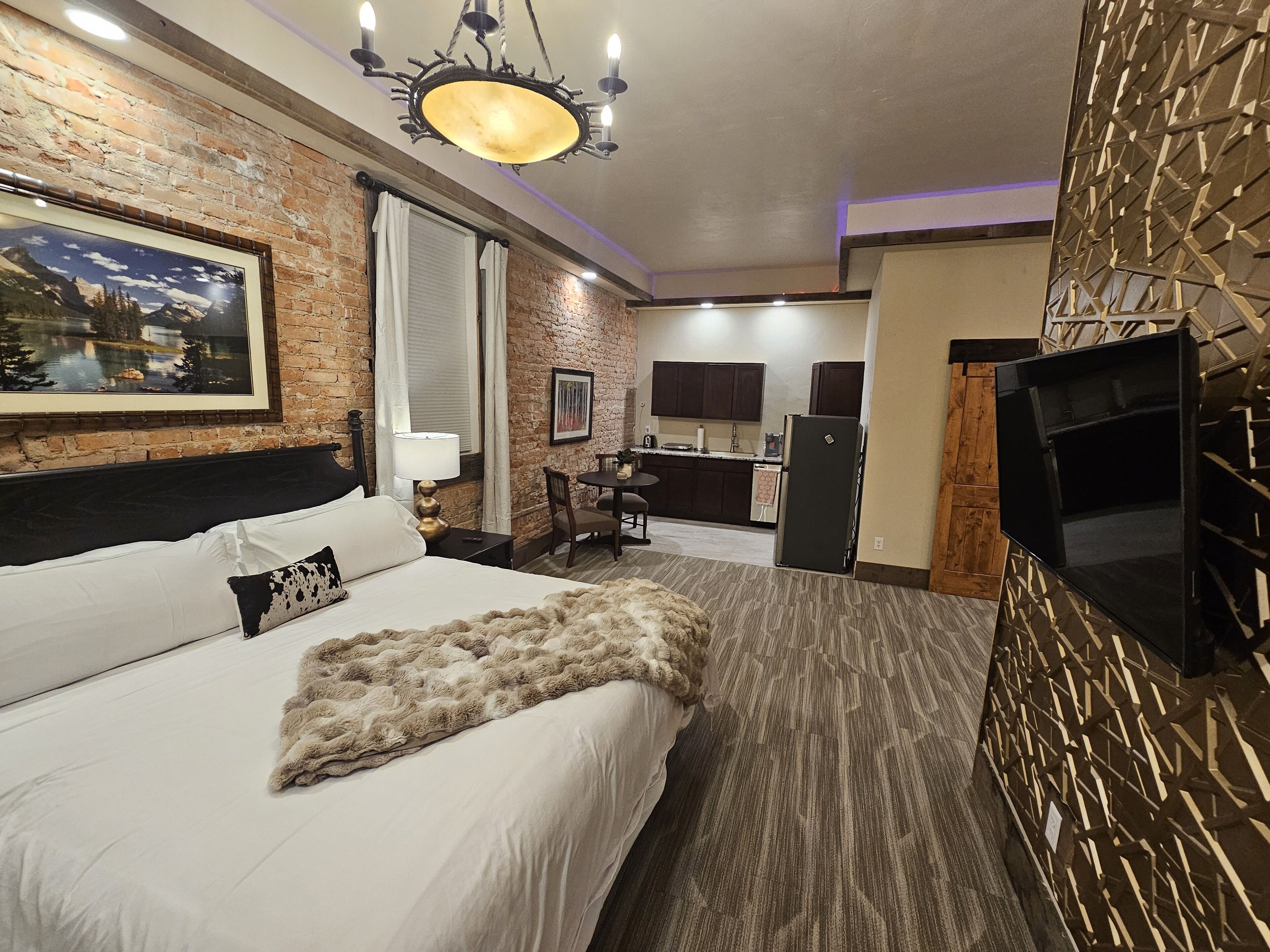 A hotel room with a bed, a desk, a chair, a TV, and a kitchen area.