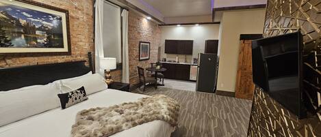A hotel room with a bed, a desk, a chair, a TV, and a kitchen area.
