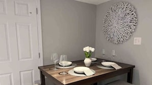 Een eetgedeelte met een houten tafel gedekt voor vier personen, witte stoelen, een vaas met witte bloemen en een decoratief wandobject.