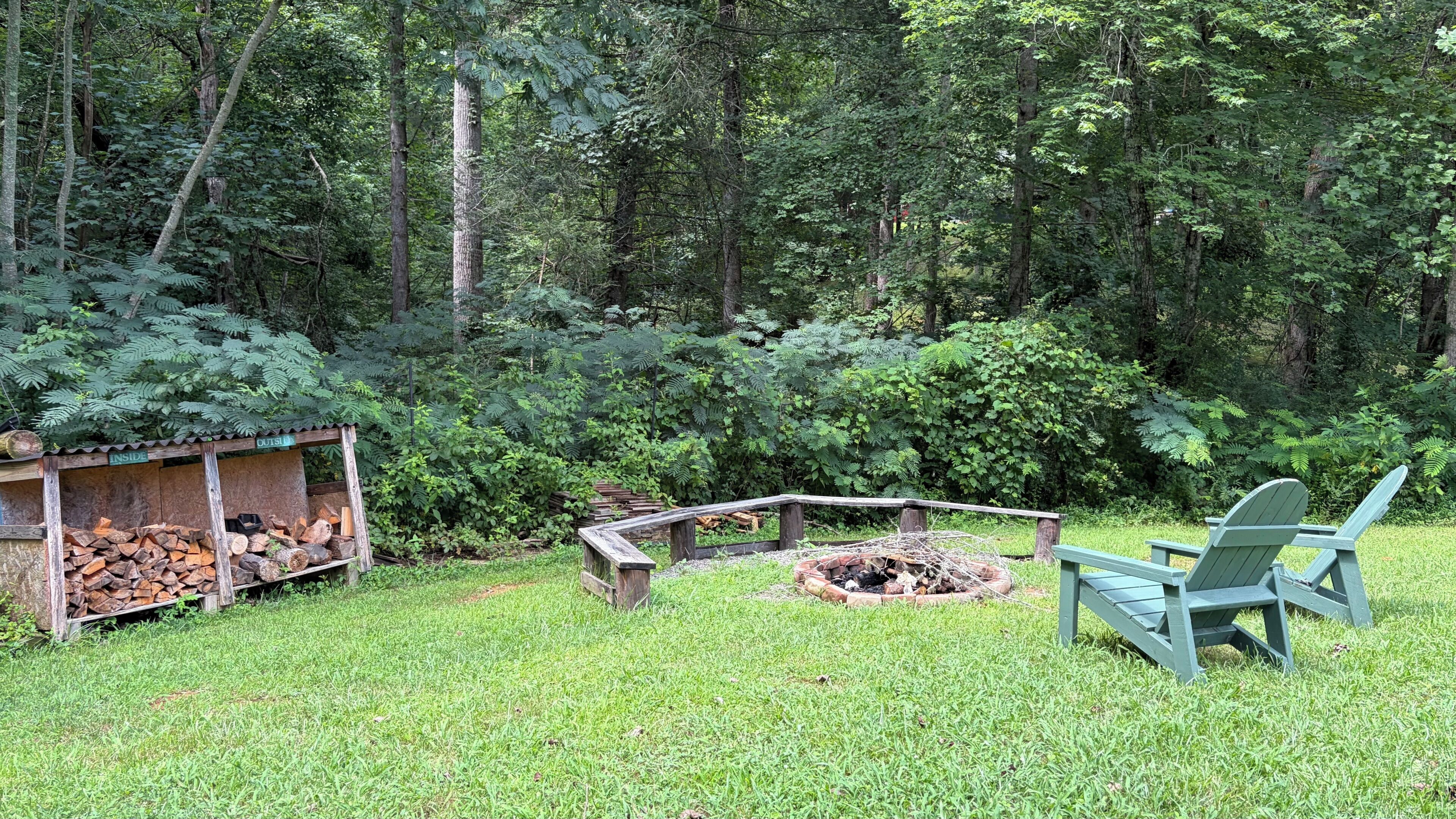 Un espace extérieur verdoyant avec un foyer, un banc en bois et une pile de bois de chauffage.