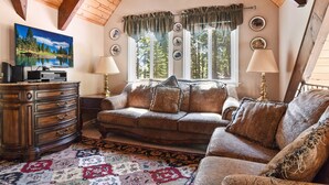 A cozy living room with a wooden ceiling, a large window with curtains, a sofa, a TV, and a wooden cabinet.