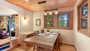 A dining area with a wooden table set for four, large windows with a view of trees, and a ceiling with wooden beams. - Lodgepole Lair - Remodeled, Walkable, Lake & Ski (Tahoe City)