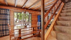 Interior de una cabaña de madera con comedor, una escalera y vistas por una ventana.