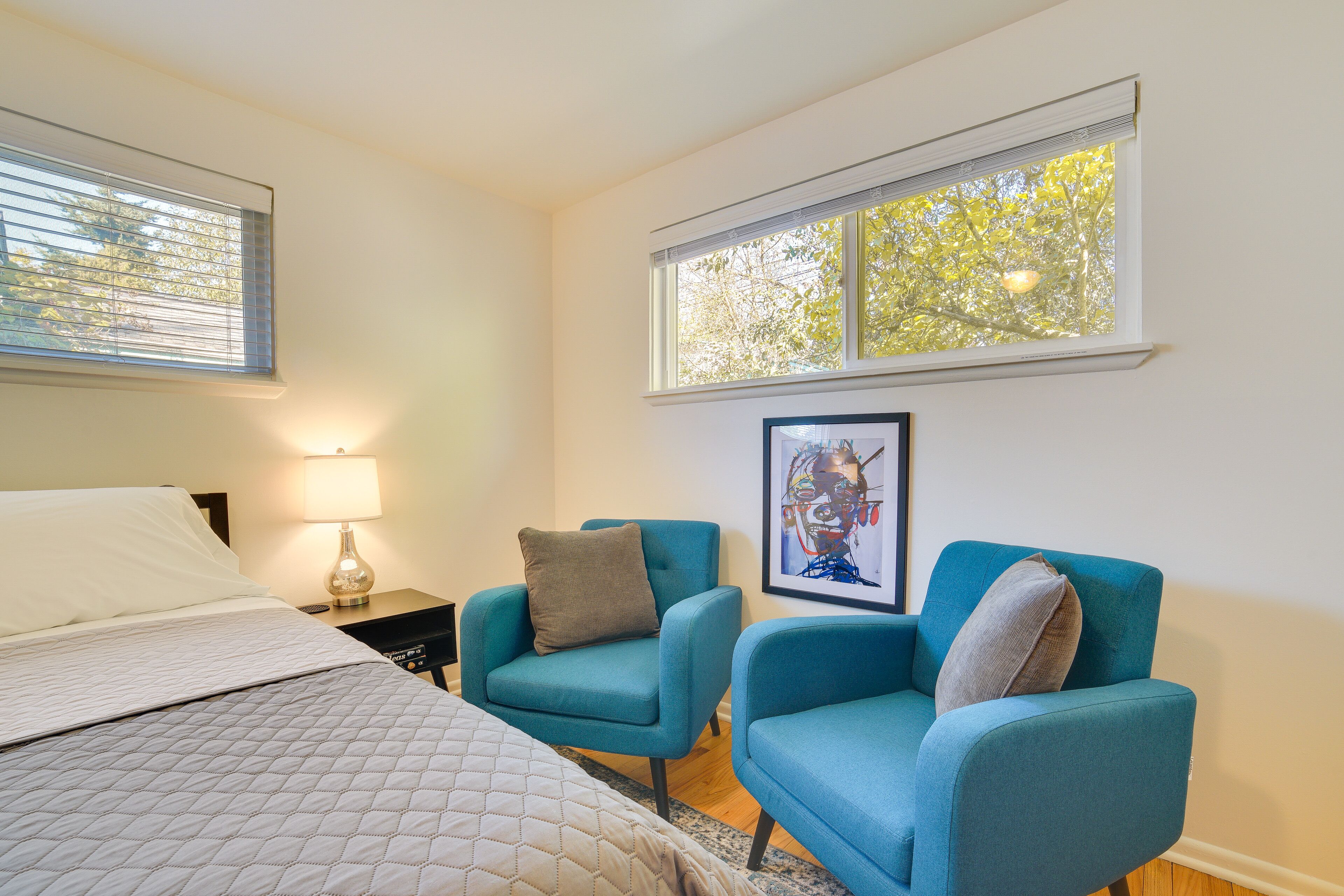 A bedroom with a bed, two blue armchairs, a nightstand with a lamp, and a framed artwork on the wall.
