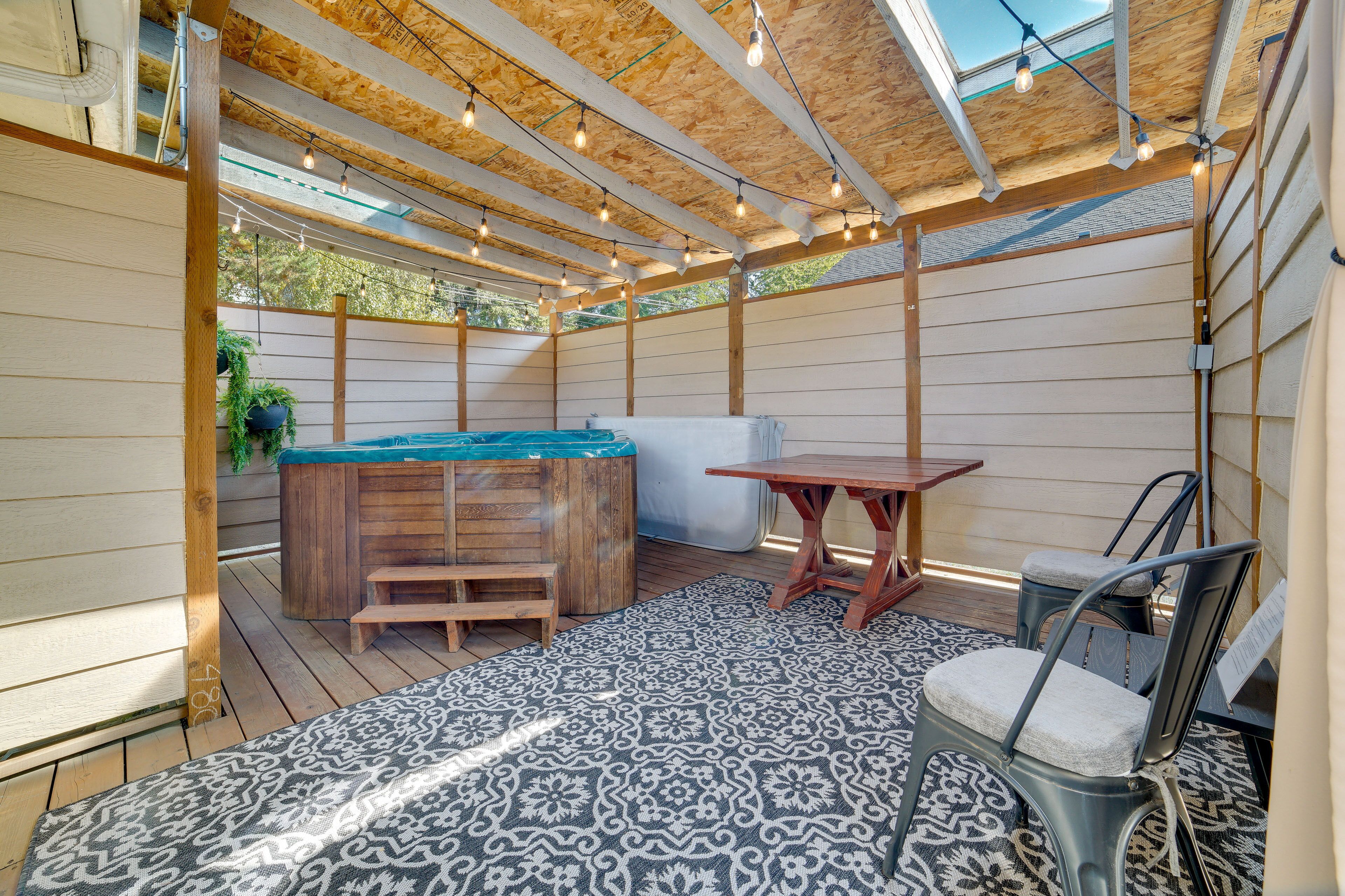 A covered outdoor area with a wooden deck, a hot tub, a table, and chairs.