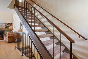 Un escalier avec des rampes en bois et des balustrades en métal, des marches recouvertes de moquette, et une vue sur un salon avec un canapé et une cheminée.