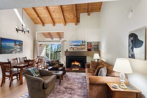 A cozy living room with a fireplace, wooden beams, a dining area, and a view of the outdoors. - Carnelian Woods Lake View Condo (Carnelian Bay)