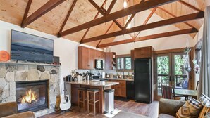 Amplia sala de estar con chimenea de piedra, vigas de madera, una cocina con una isla grande y un comedor con vistas al exterior.