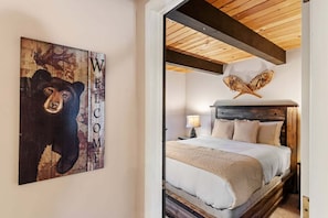 Un dormitorio con cabecera de madera, cama con ropa blanca, lámpara en una mesita de noche y un cartel de 'WELCOME' con una ilustración de un oso.