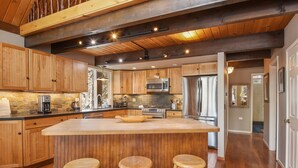 A spacious kitchen with wooden cabinets, a large island with stools, a stainless steel refrigerator, and a stone backsplash.