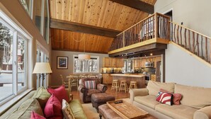 A spacious living area with a high ceiling, wooden beams, a dining area, and a kitchen with a bar.