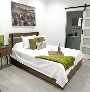 A neatly arranged bedroom with a bed, pillows, a bedside table, a tray with glasses and a bottle, and a cityscape painting on the wall.