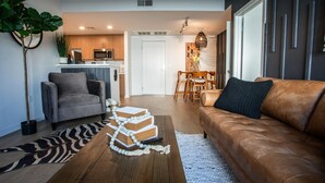 Ein modernes Wohnzimmer mit einer Couch, einem Couchtisch mit Büchern und einer Fernbedienung, einem Essbereich mit Tisch und Stühlen sowie einer Küche im Hintergrund.