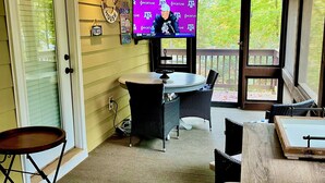 Un espacio al aire libre techado con una televisión mostrando un noticiero, muebles de mimbre y una mesa de madera.