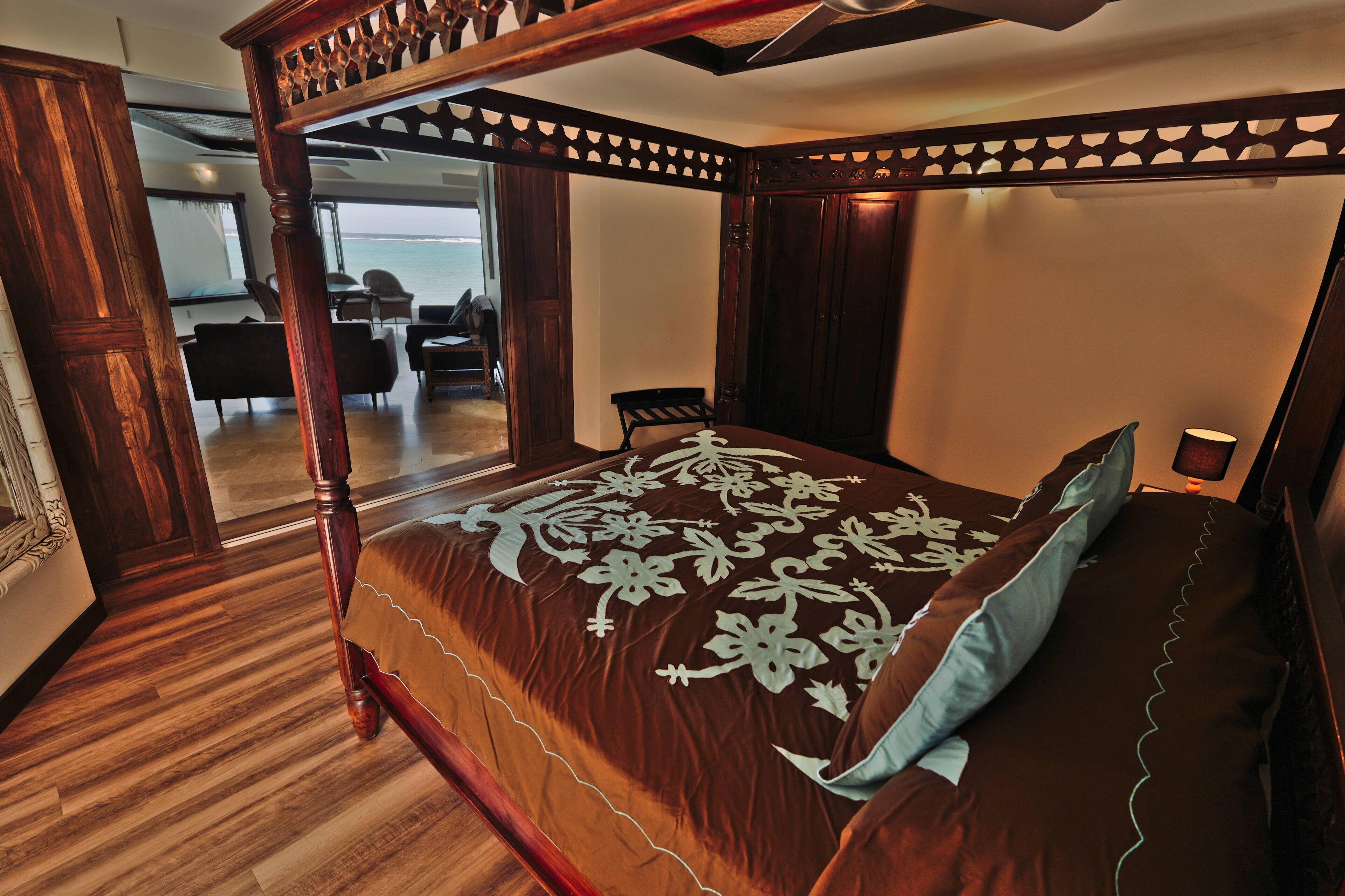 A four-poster bed with a floral bedspread, a wooden floor, and a view of the sea through an open door.