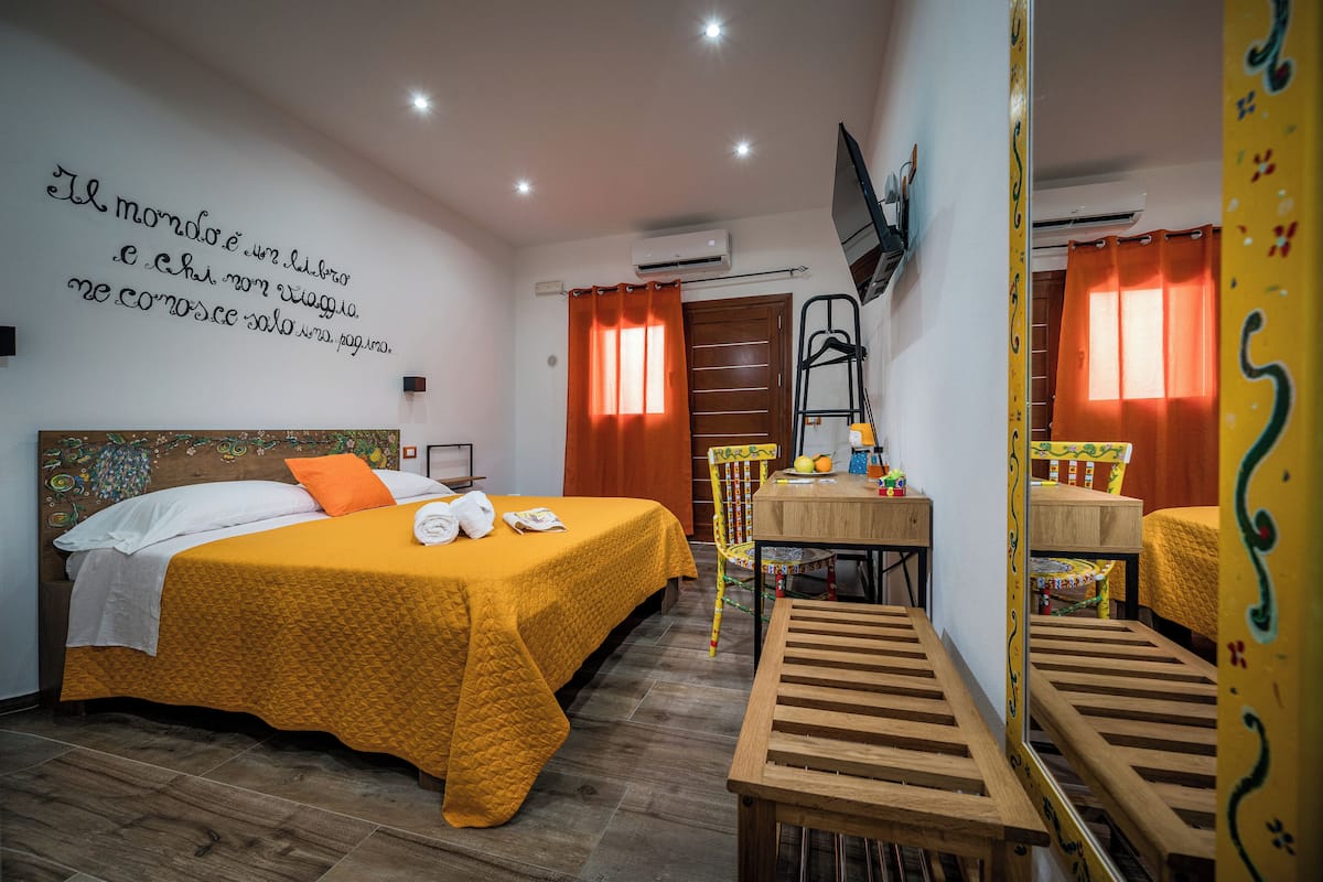 A hotel room with a bed, a desk, and a chair. There is a wall with a quote, a TV mounted on the wall, and a window with curtains.