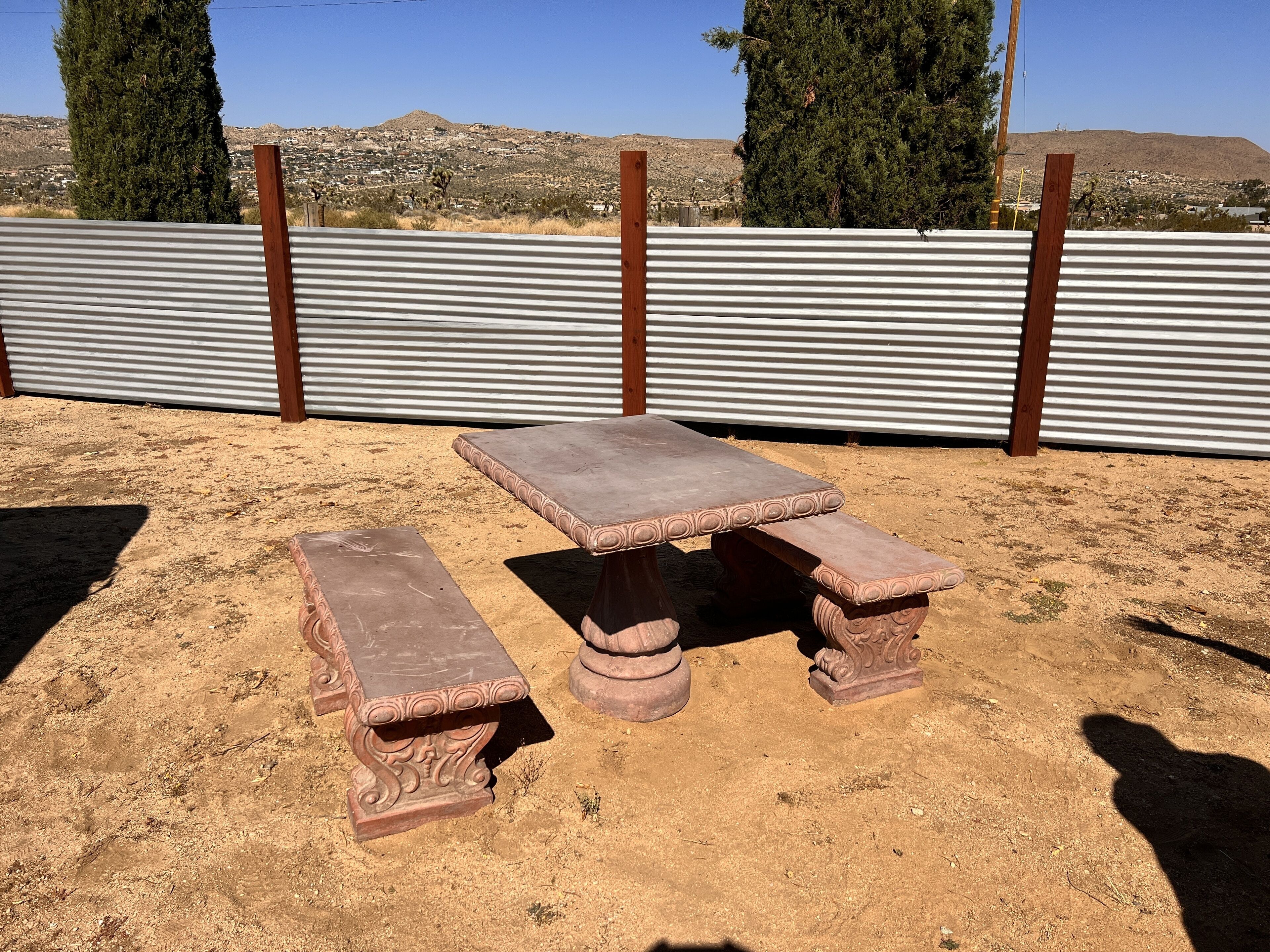 Un espace extérieur rustique aménagé avec une table et des bancs en pierre, entouré d’une clôture en métal ondulé et offrant en arrière-plan un paysage désertique.
