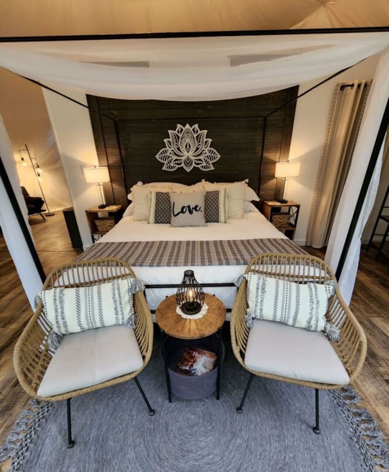 A spacious bedroom with a large bed, two bedside tables with lamps, a decorative wall piece, and two wicker chairs with cushions.